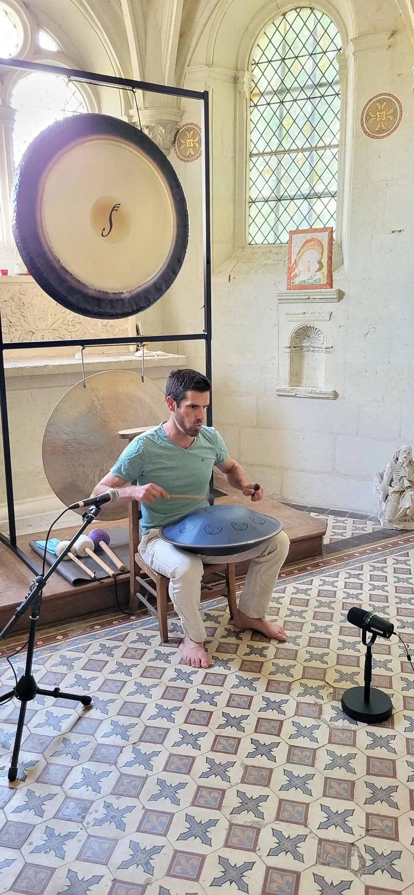Alexandre Tonnelier jouant du handpan devant deux gongs dans une chapelle aux murs de pierre claire