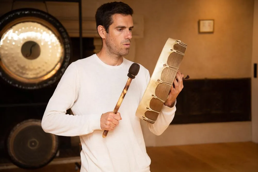 Alexandre Tonnelier tenant un tambour chamanique devant deux gongs